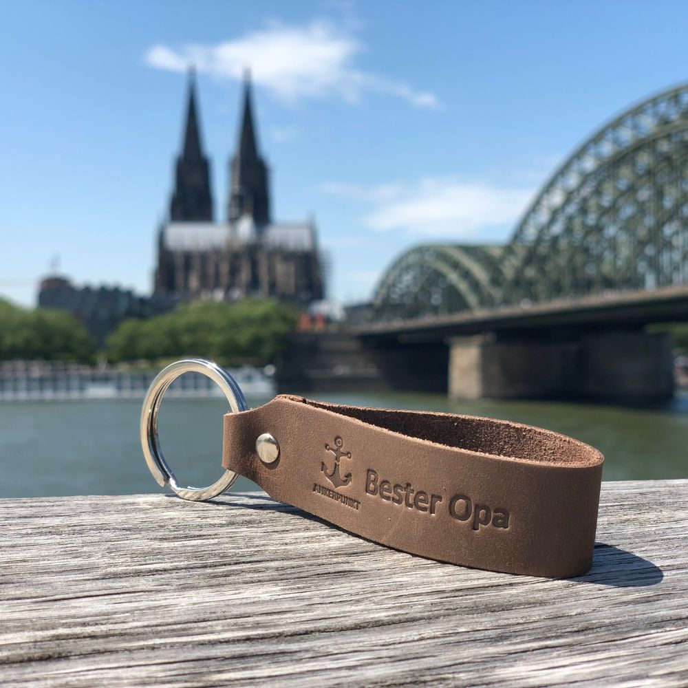 Schlüsselanhänger mit Gravur Bester Opa in dunkelbraun liegt auf Holz am Rhein, im Hintergrund ist der Kölner Dom zu sehen