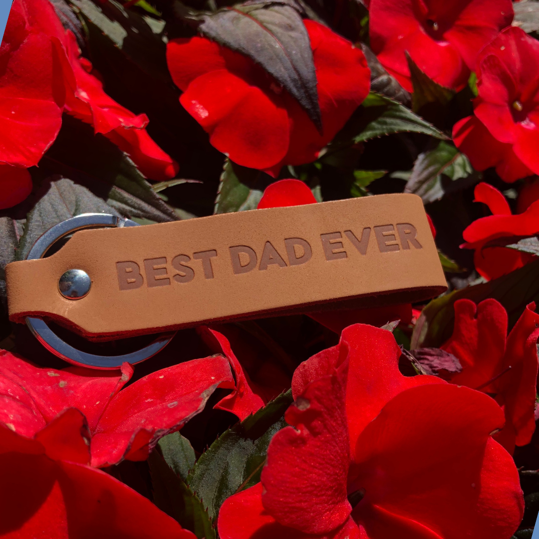 Ankerpunkt Schlüsselanhänger aus Leder mit Gravur Best Dad Ever hellbraun auf roten Blumen im Park.