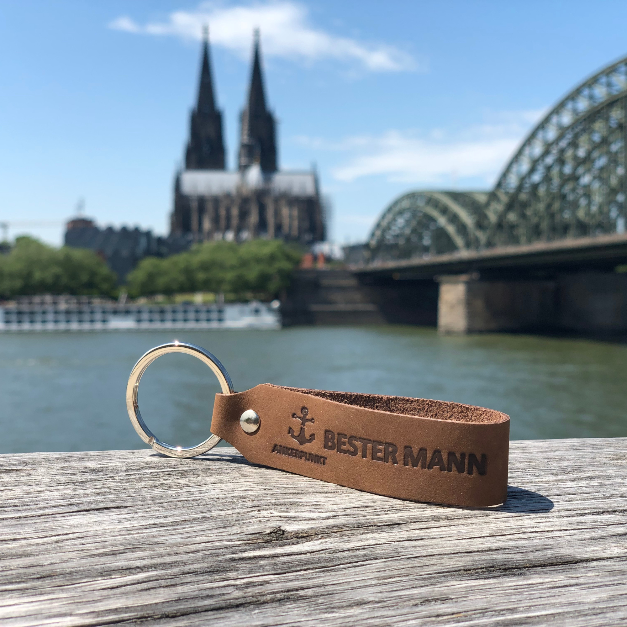 Schlüsselanhänger mit Gravur Bester Mann in dunkelbraun liegt auf Holz am Rhein, im Hintergrund ist der Kölner Dom zu sehen