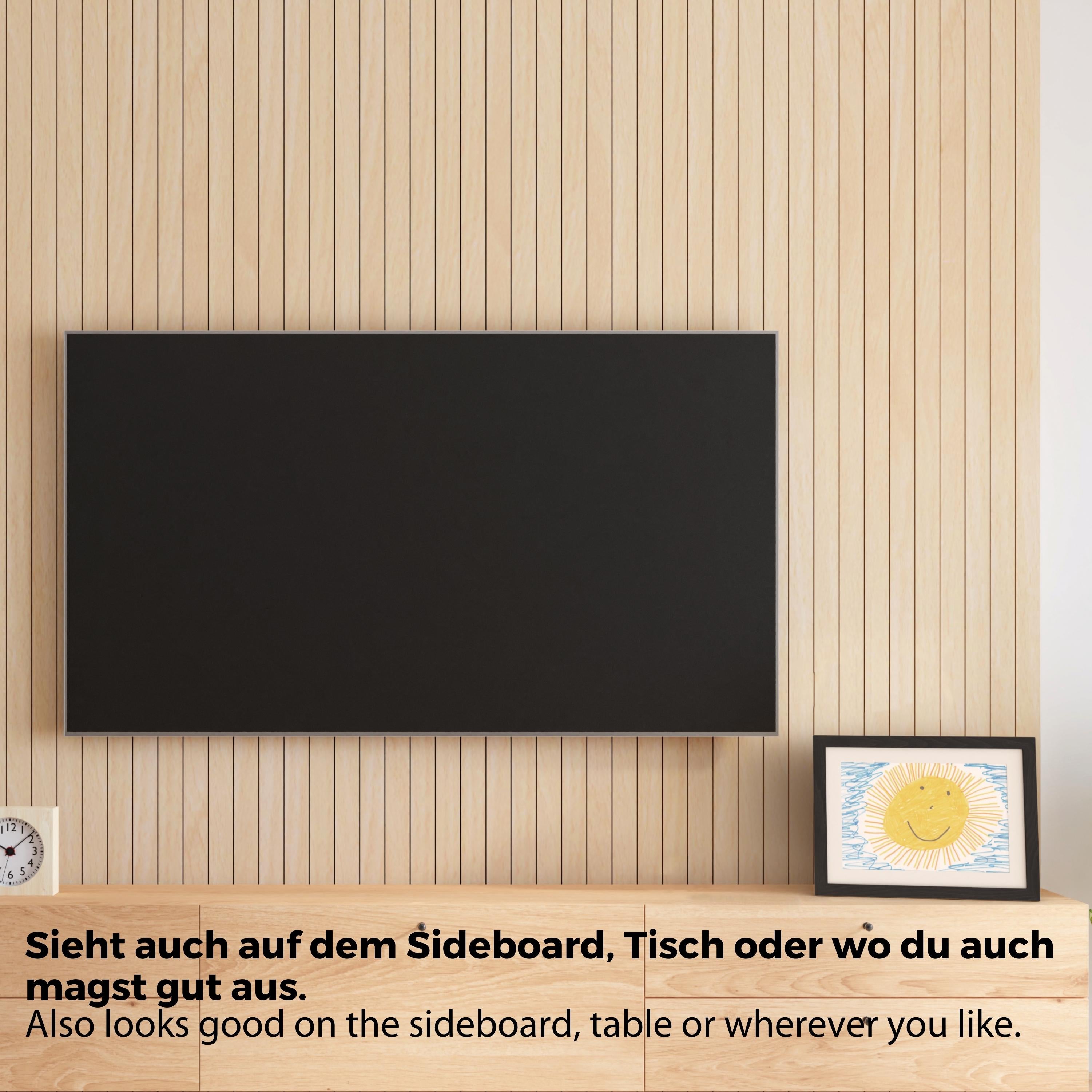 Schwarzer Bilderrahmen mit Bild steht auf einer Fernsehbank. Neben dem Rahmen hängt ein Fernseher an der Wand.