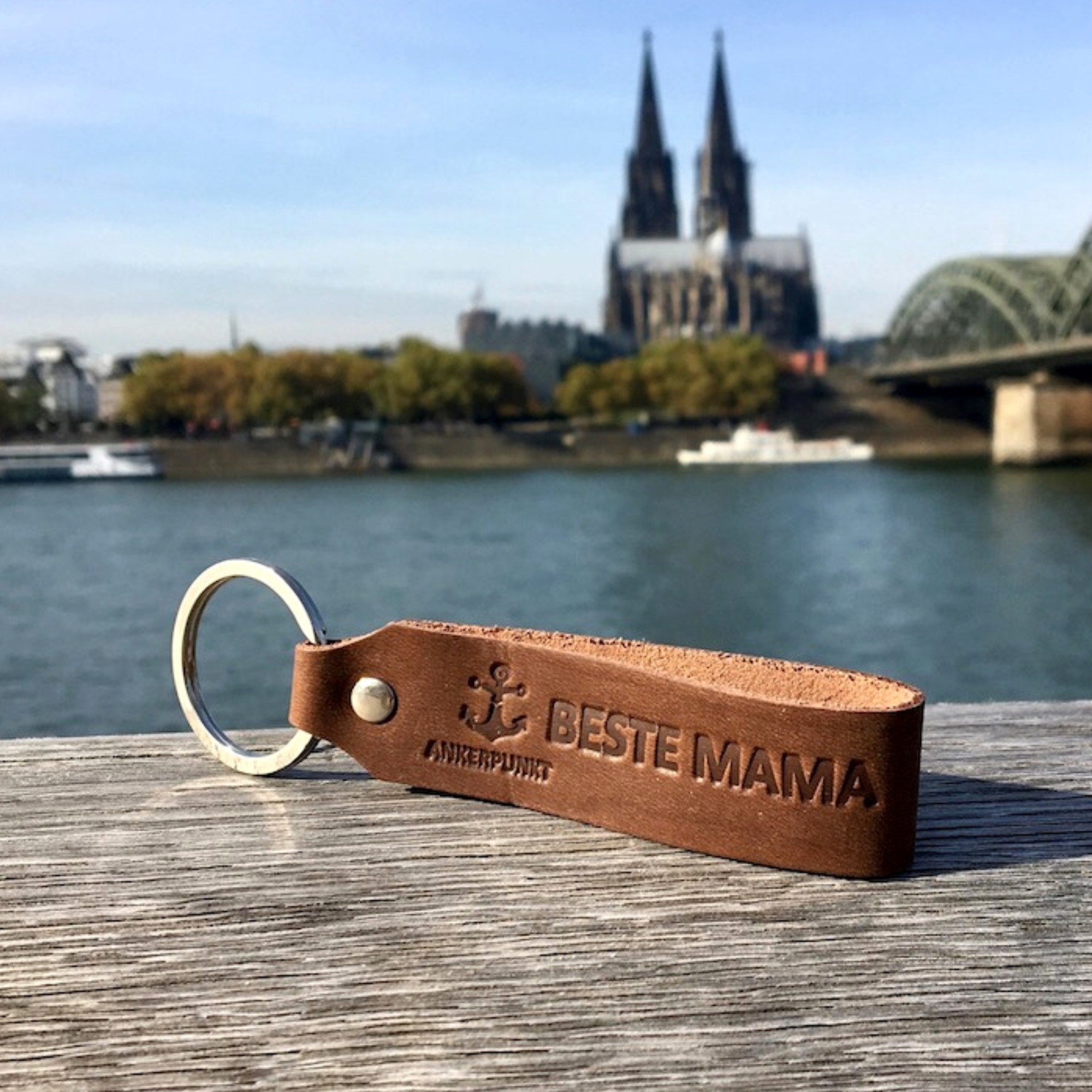 Beste Mama Schlüsselanhänger dunkelbraun liegt auf Holz am Rhein, im Hintergrund ist der Kölner Dom zu sehen