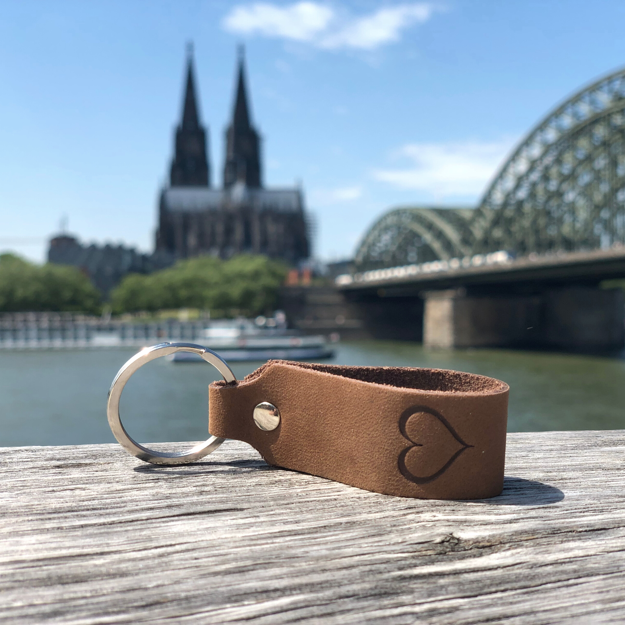 Schlüsselanhänger mit Gravur Herz liegt auf Holz am Rhein, im Hintergrund ist der Kölner Dom zu sehen